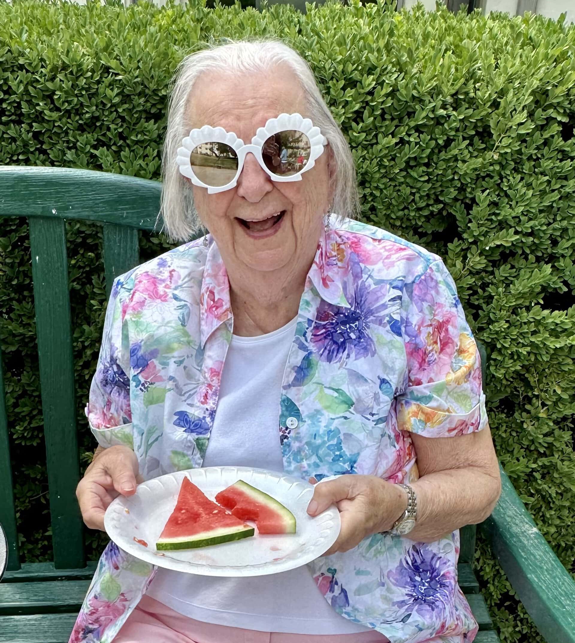 happy senior lady outside sitting on a green bench holding two slices of watermelon