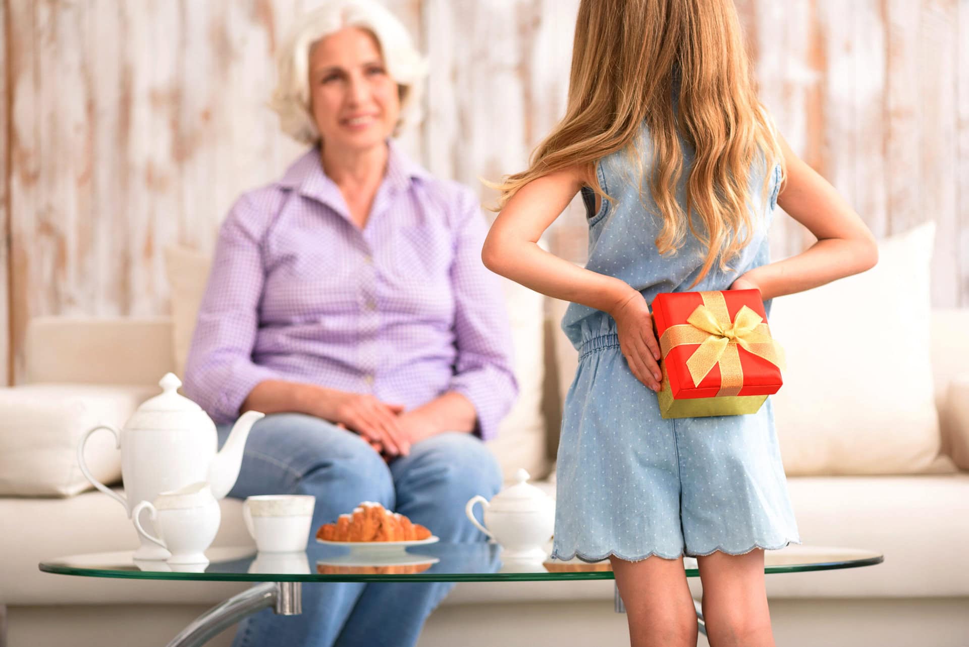 a little girl with a present behind her back to surprise her grandmother sitting on a white couch